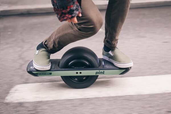Person riding olive green onewheel pint board down a street.